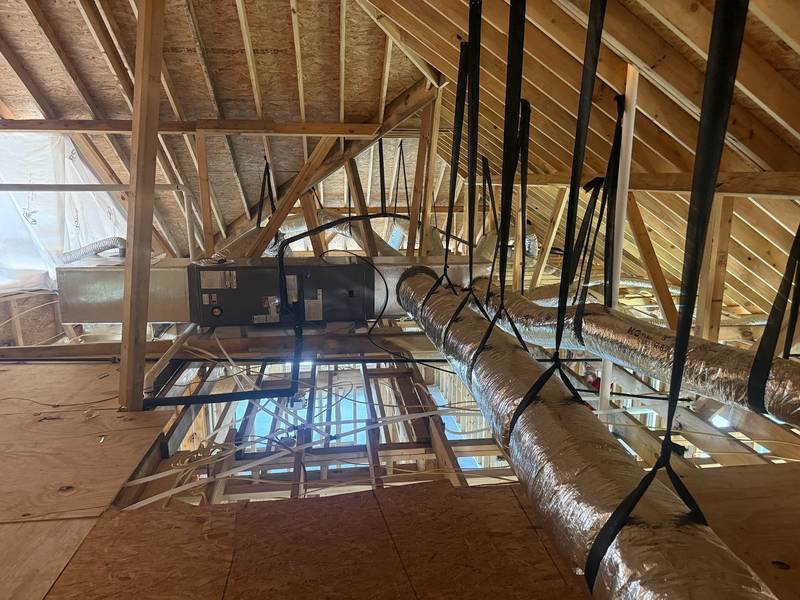 Looking up into an open-framed attic at an air handler on a platform, with silver insulated flex ducts hung on black straps
