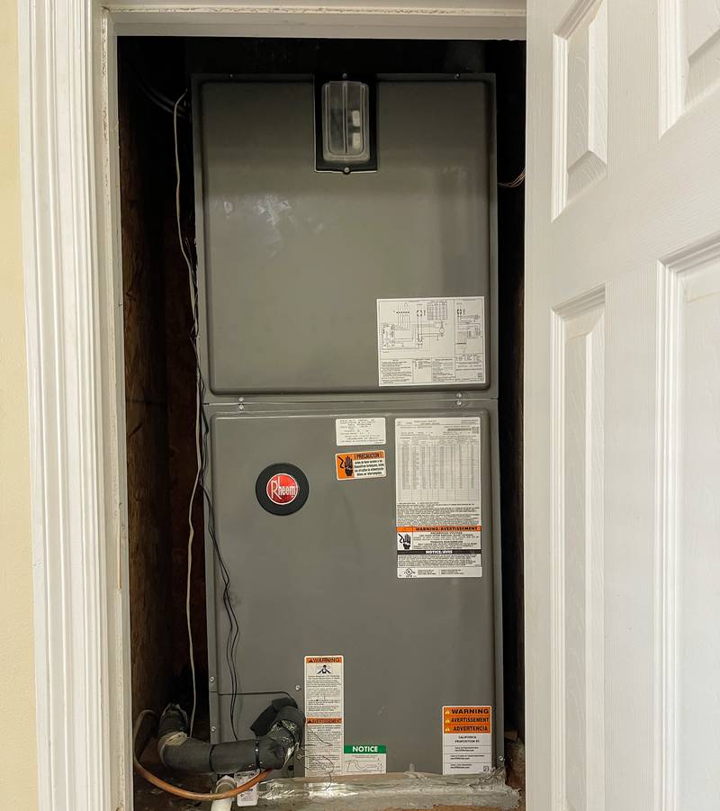 Gray Rheem upflow air handler standing in a hall closet with the white door open beside it