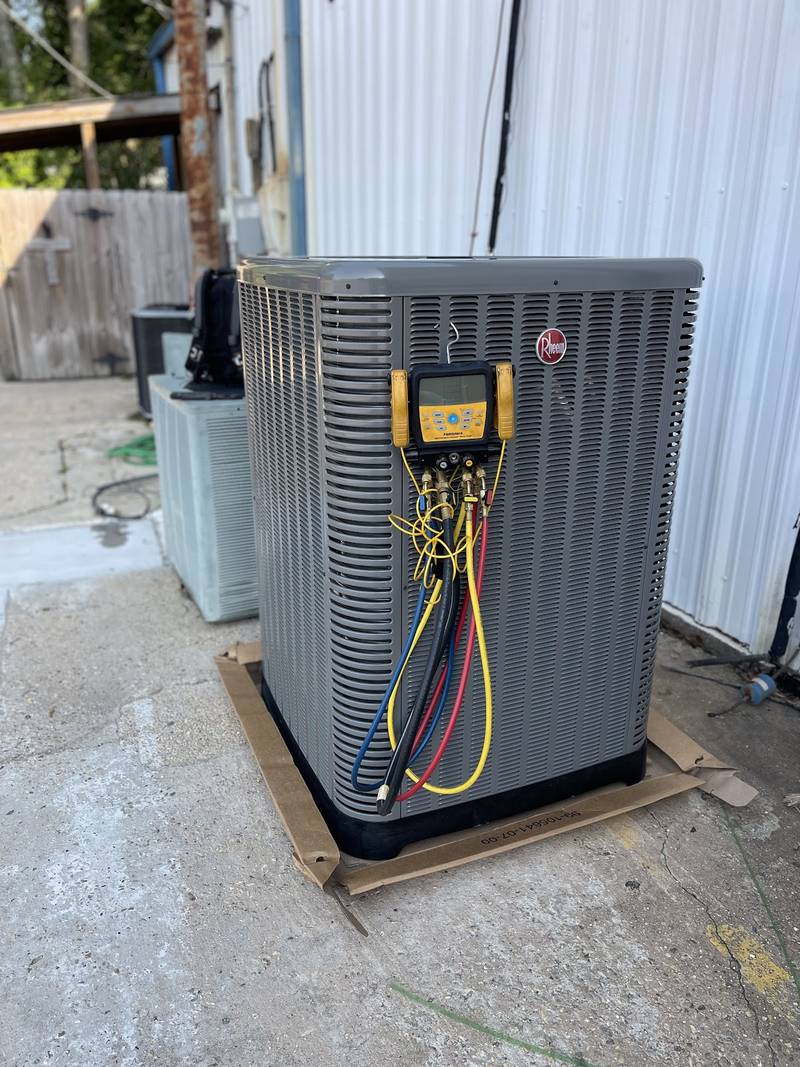 New Rheem condenser on its shipping cardboard on a concrete pad beside a white metal building, with a Fieldpiece digital manifold and red, blue and yellow hoses hanging on it