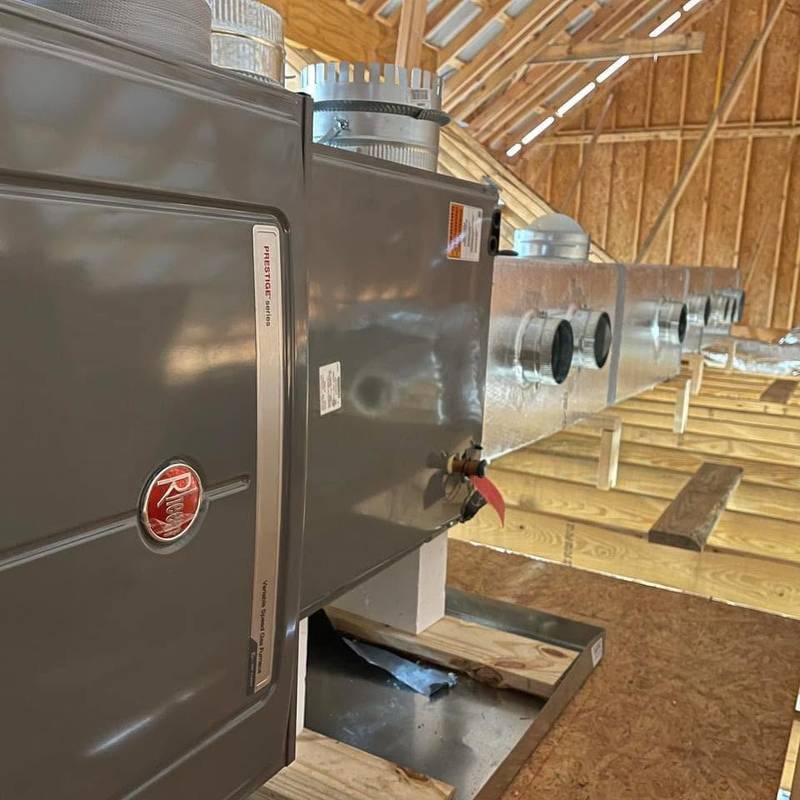Gray Rheem Prestige series gas furnace in an attic, with a supply plenum and round duct collars running along the floor joists