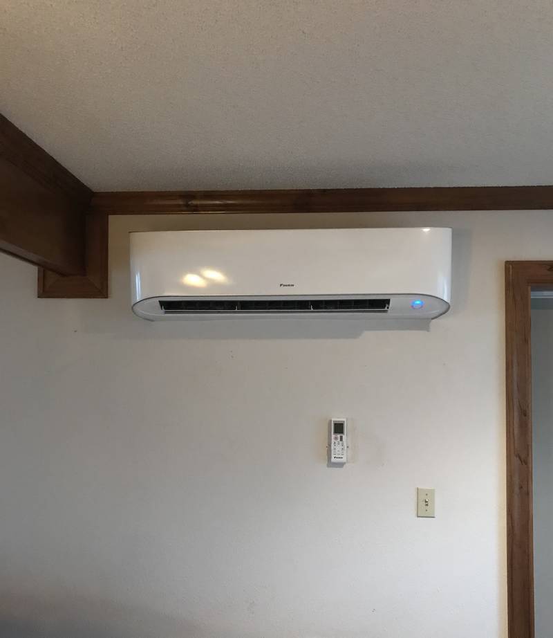 White Daikin ductless wall unit mounted high on a white wall under wood trim, with a remote holder below