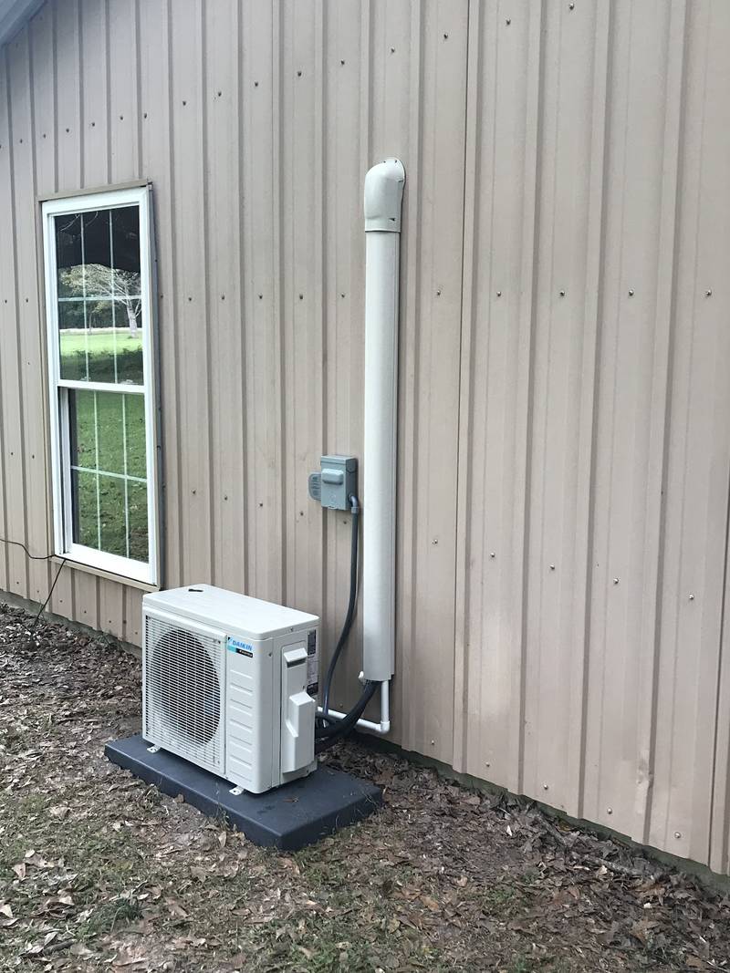 Daikin mini-split outdoor unit on a black pad beside a tan metal-sided building, with a white line-set cover running up the wall