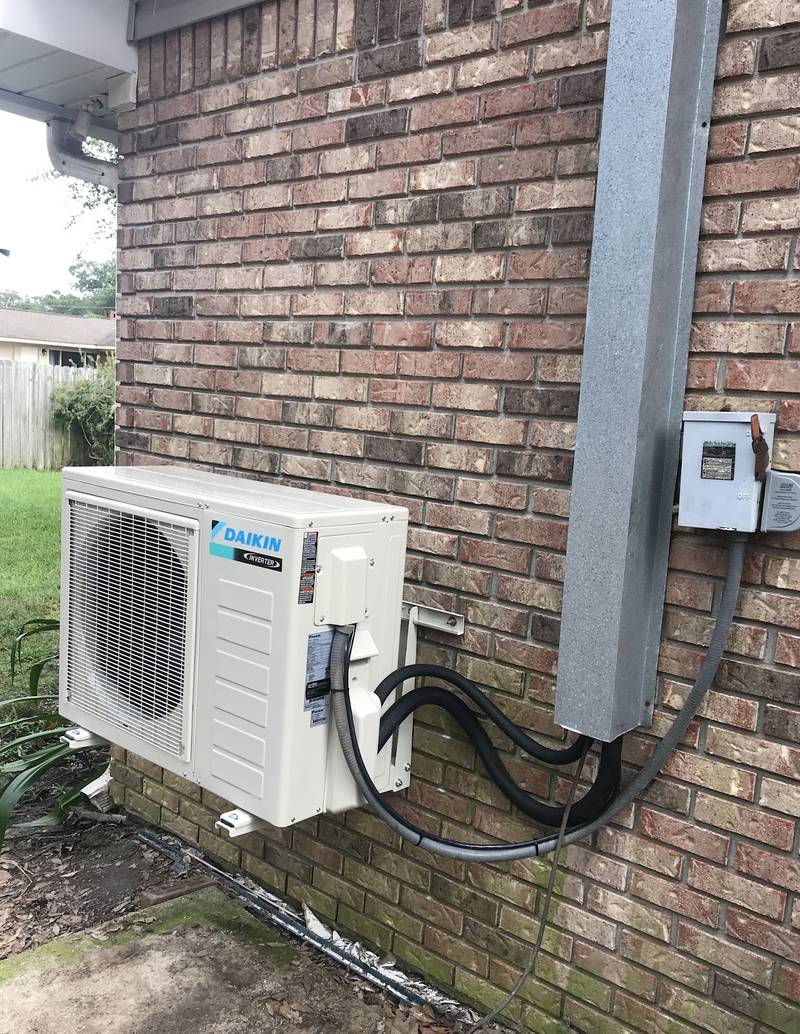 Daikin Inverter mini-split outdoor unit on a wall bracket against a brick house, with the line set and disconnect box beside a downspout